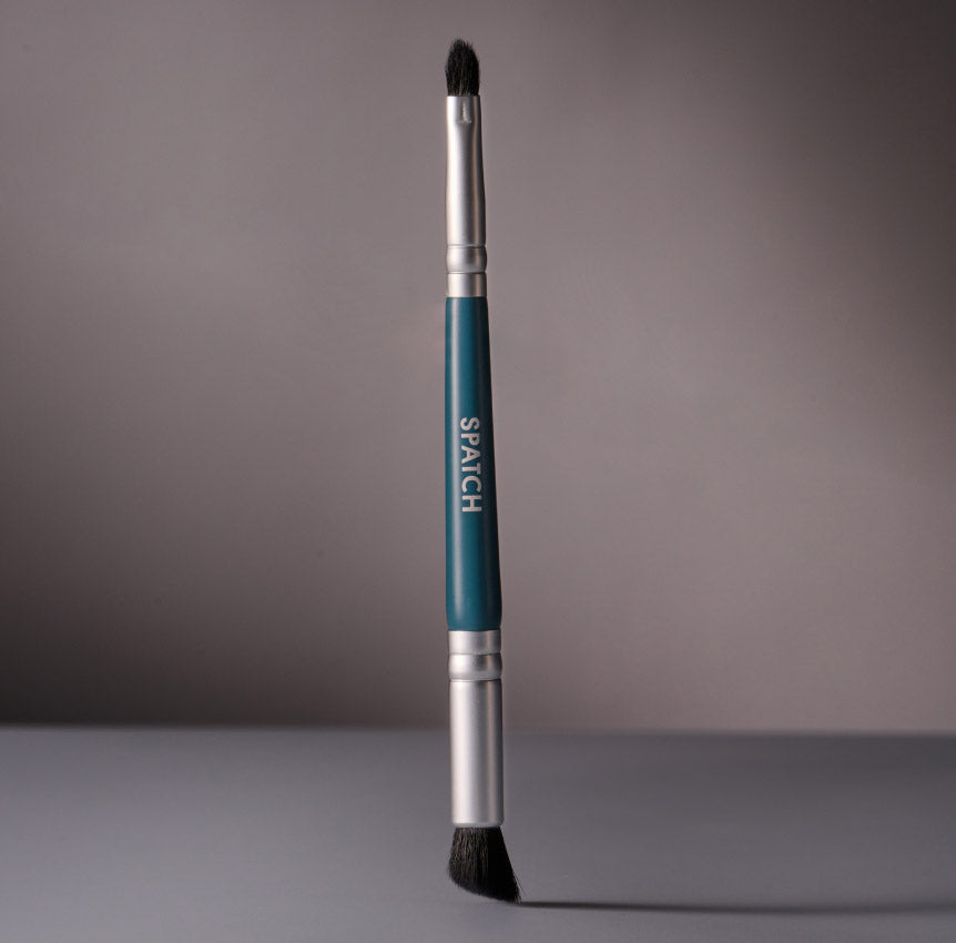 A double-sided teal-colored makeup brush balances upright on a gray background