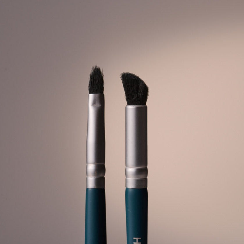 Two makeup brushes with dark teal handles and silver ferrules stand against a softly lit gradient background