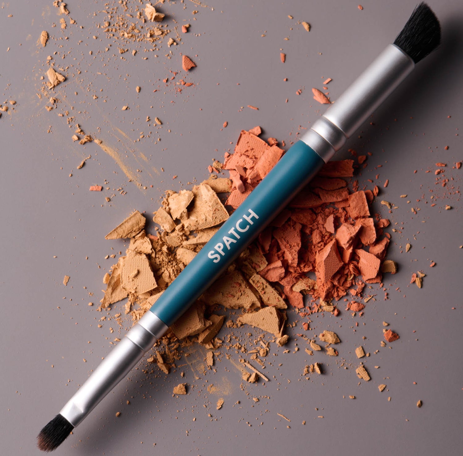 A double sided teal colored makeup brush sits on a pile of broken peach-colored pressed powder makeup