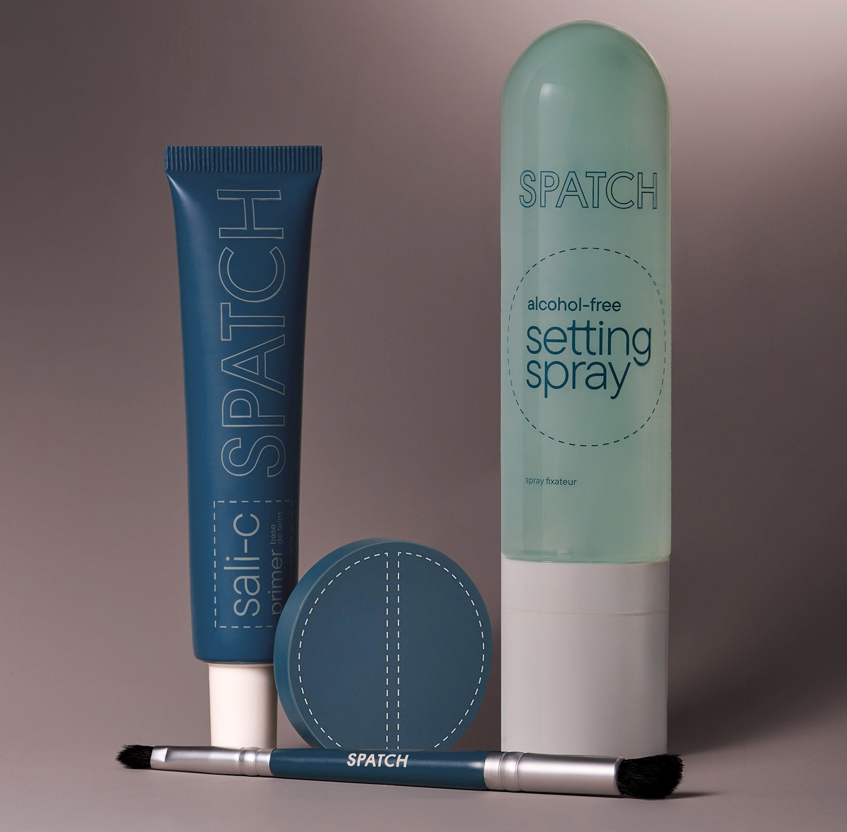 a group of four cosmetic products on a gray background. From left to right: a teal colored tube of SPATCH Sali-C primer, a teal colored makeup brush, a round, teal-colored pressed powder compact open to show two shades of tan-colored powder, and an aqua-colored bottle of SPATCH alchohol-free setting spray with a white cap