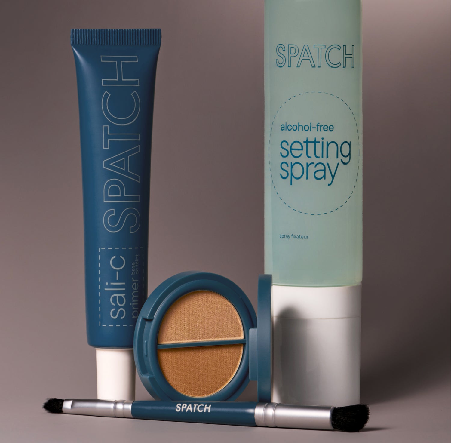 a group of four cosmetic products on a gray background. From left to right: a teal colored tube of SPATCH Sali-C primer, a teal colored makeup brush, a round, teal-colored pressed powder compact open to show two shades of tan-colored powder, and an aqua-colored bottle of SPATCH alchohol-free setting spray with a white cap