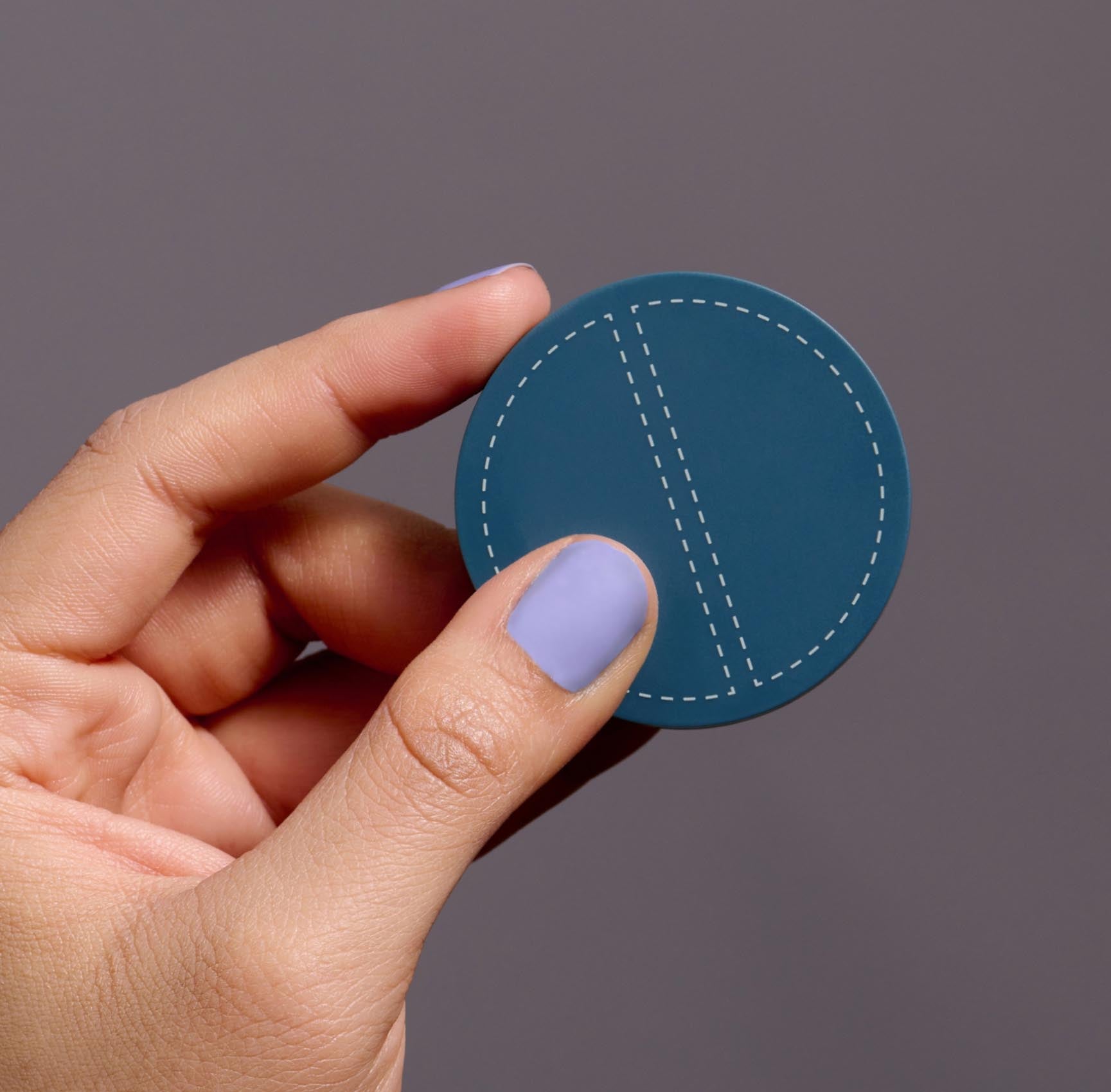 A hand with light purple nail polish holds a closed SPATCH invisible spot fix compact up against a gray background. The compact is teal-colored, circular, and about 2 inches in diameter. The compact has a graphic of two half-circles with dashed outlines on it.