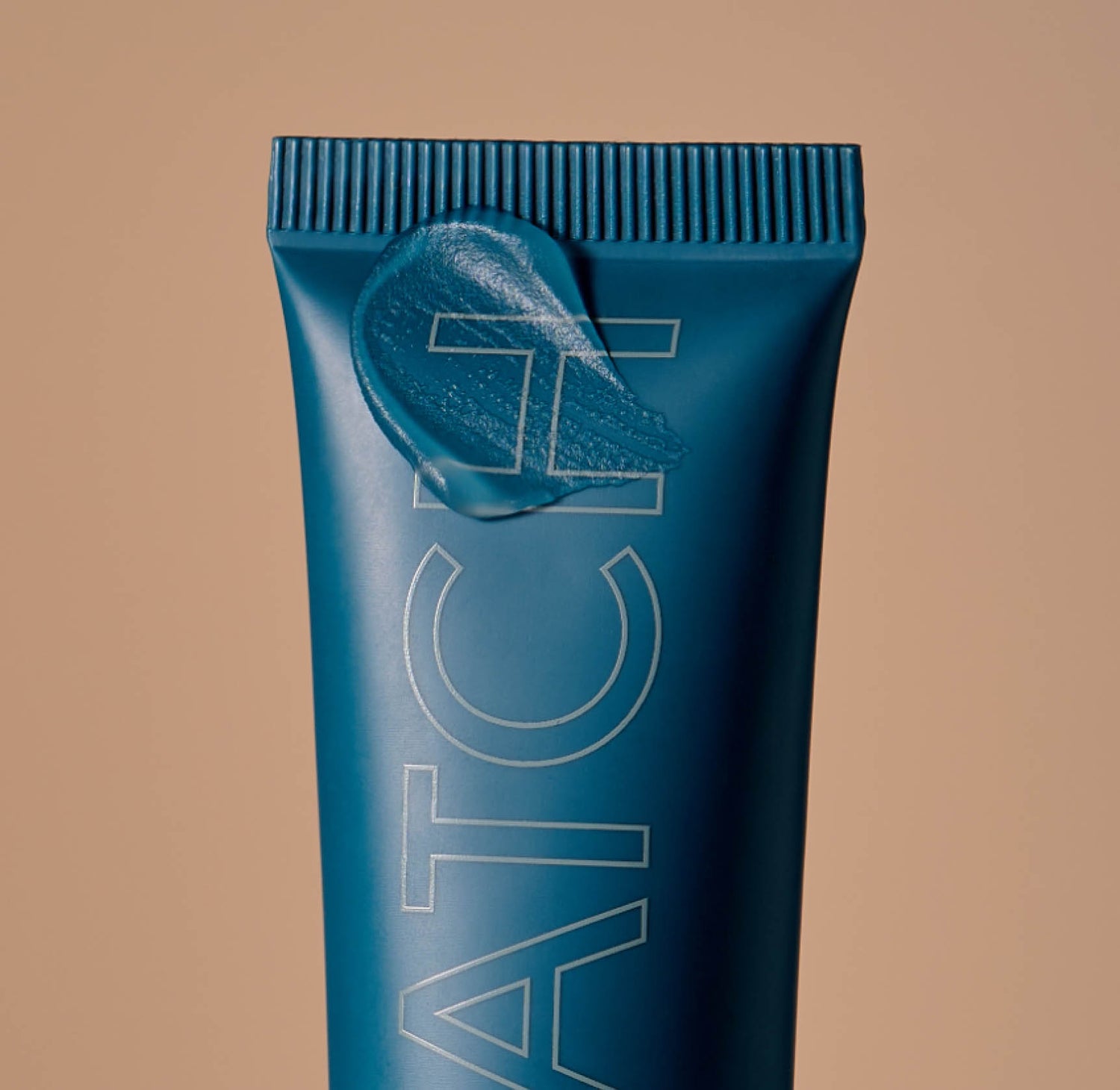 A close up photo of a teal-colored tube of makeup primer on a tan background with a blob of primer on the end of the tube