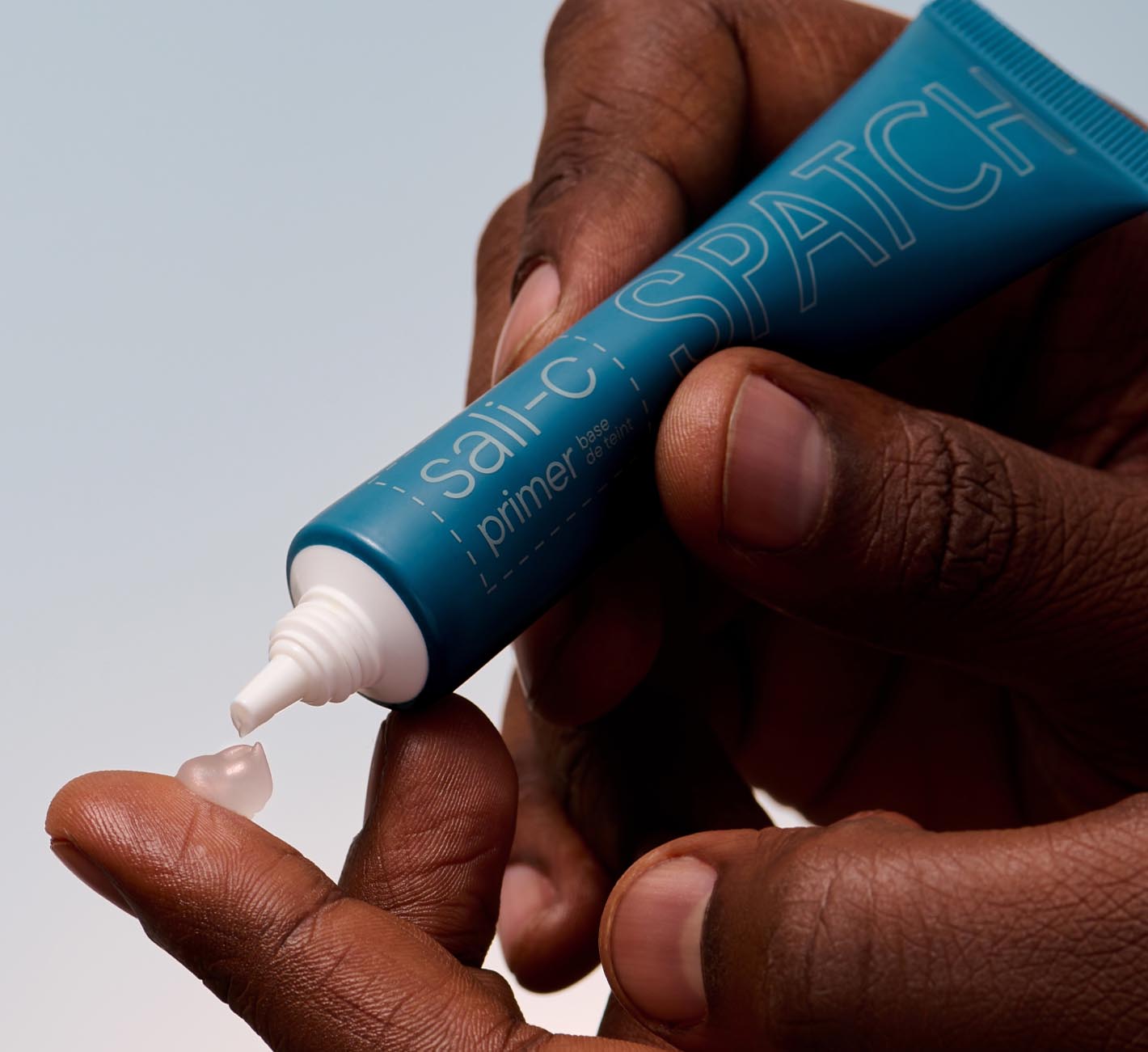 Hands hold a teal-colored tube of primer and squeeze a dollop of semi-opaque gel primer onto finger