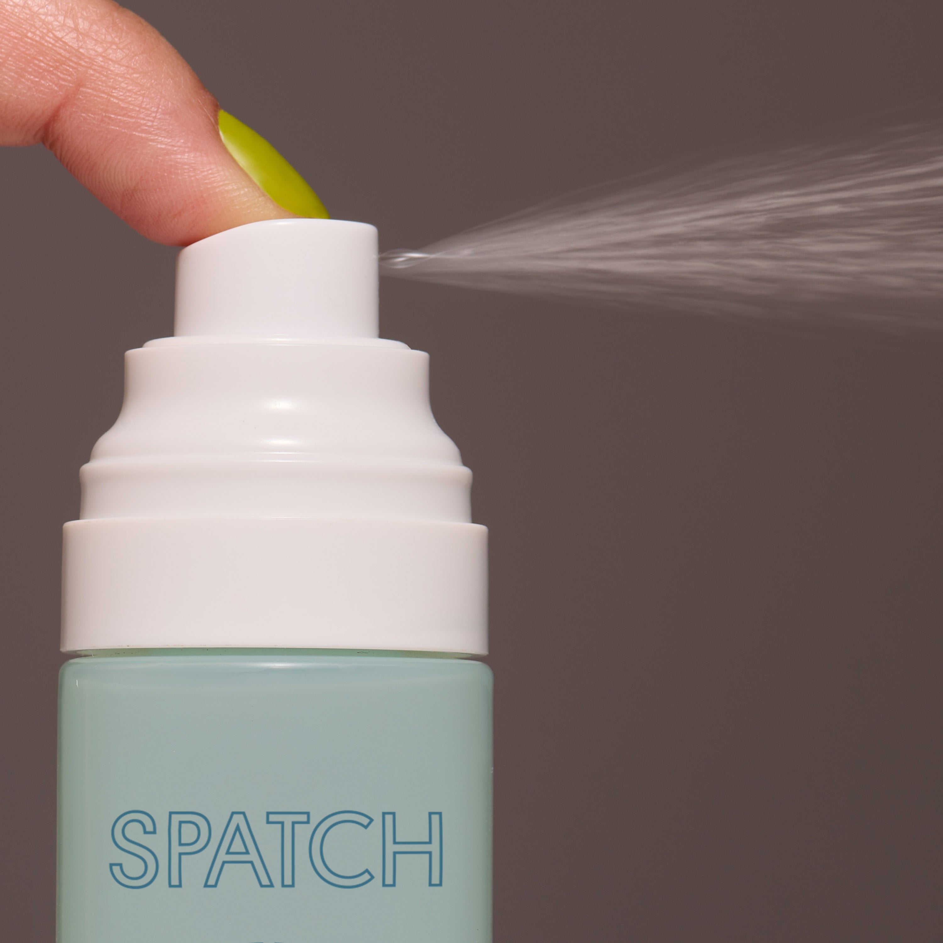 Aqua green bottle of liquid setting spray with a white nozzle spraying liquid, being pushed by a finger with yellow nail polish, on a gray background