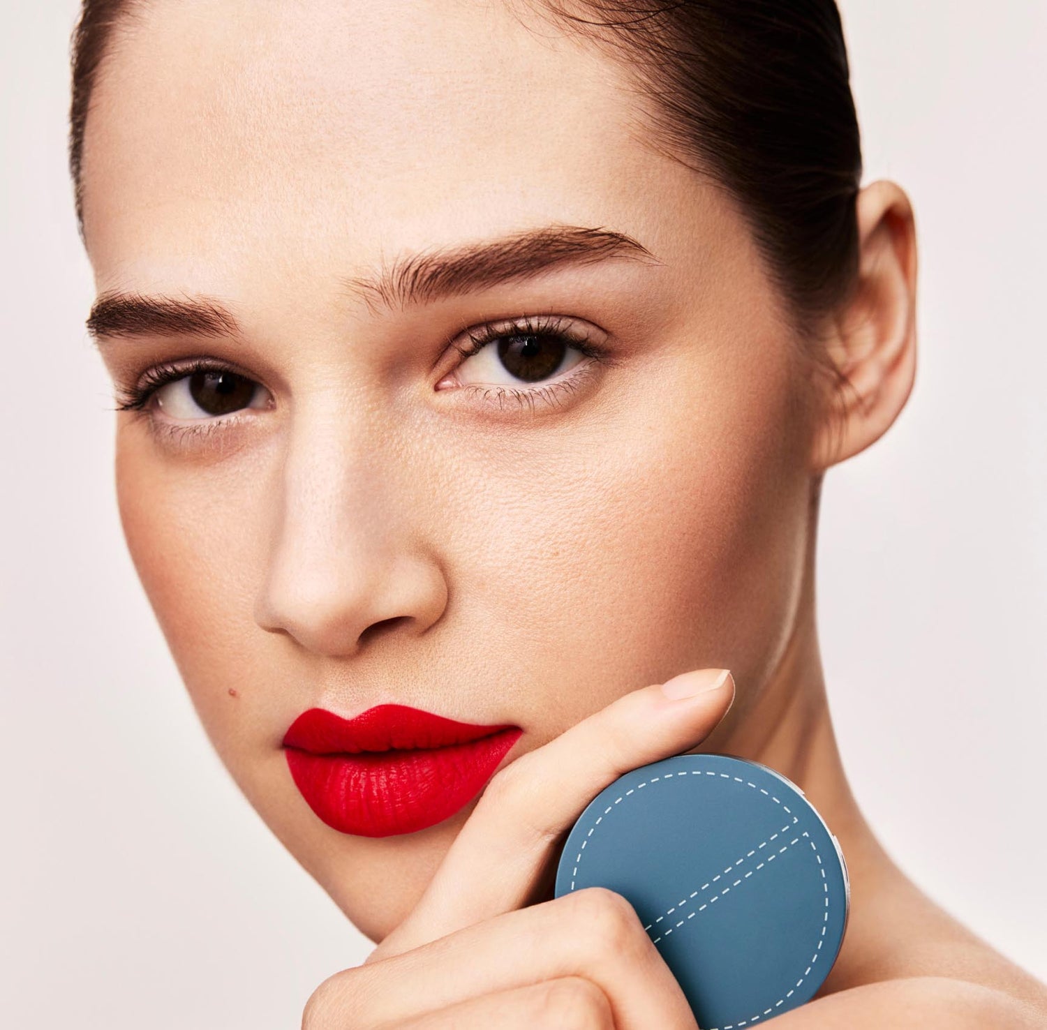 A woman with red lipstick and slicked back hair on a light gray background holds a circular, teal-colored makeup compact of SPATCH invisible spot fix up to her shoulder in the foreground of the image, covering her lower jaw. She is looking at the camera.