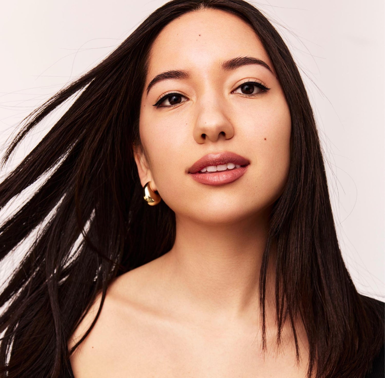 A young woman with long black hair and a gold hoop earring looks at the camera on a light gray background 