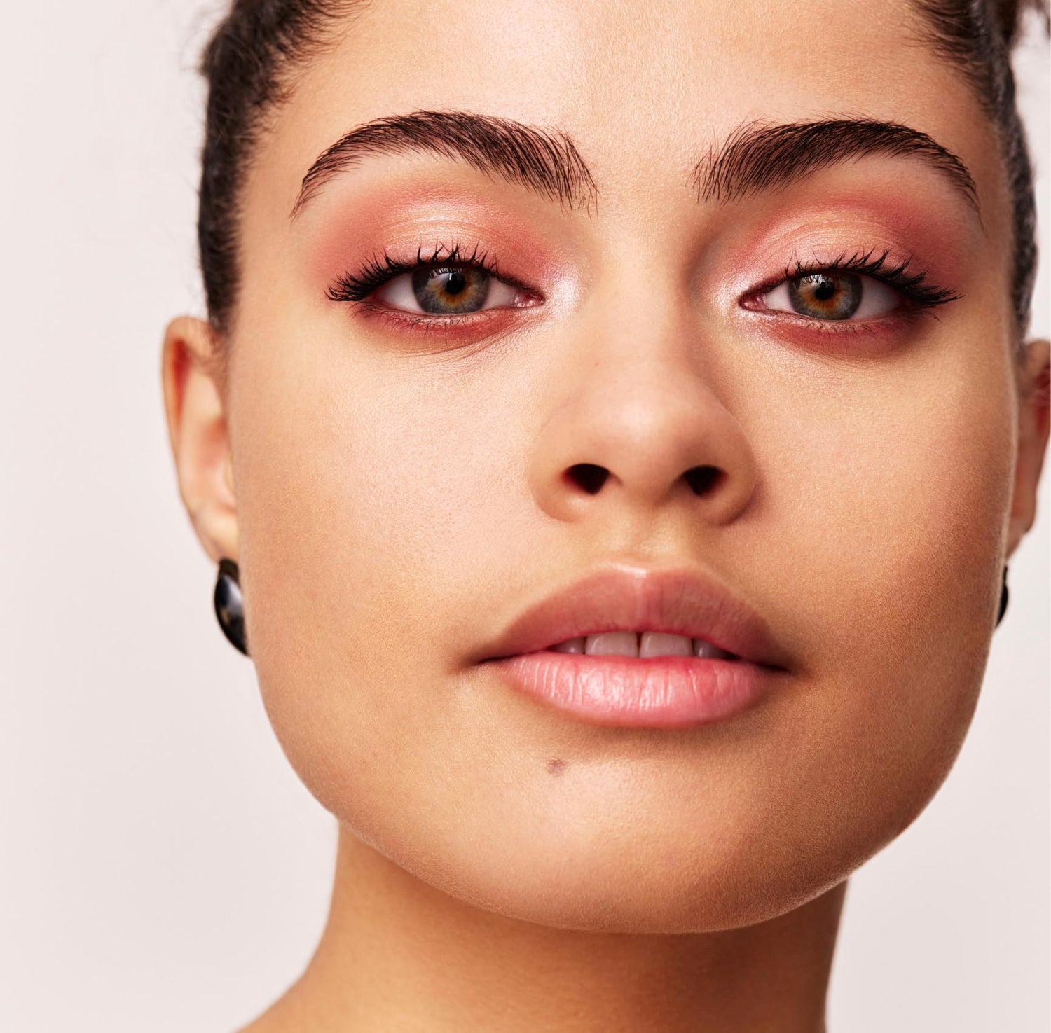 A close up image of a woman looking at the camera on a light gray background. She wears earrings and pink eyeshadow and her hair is slicked back, 