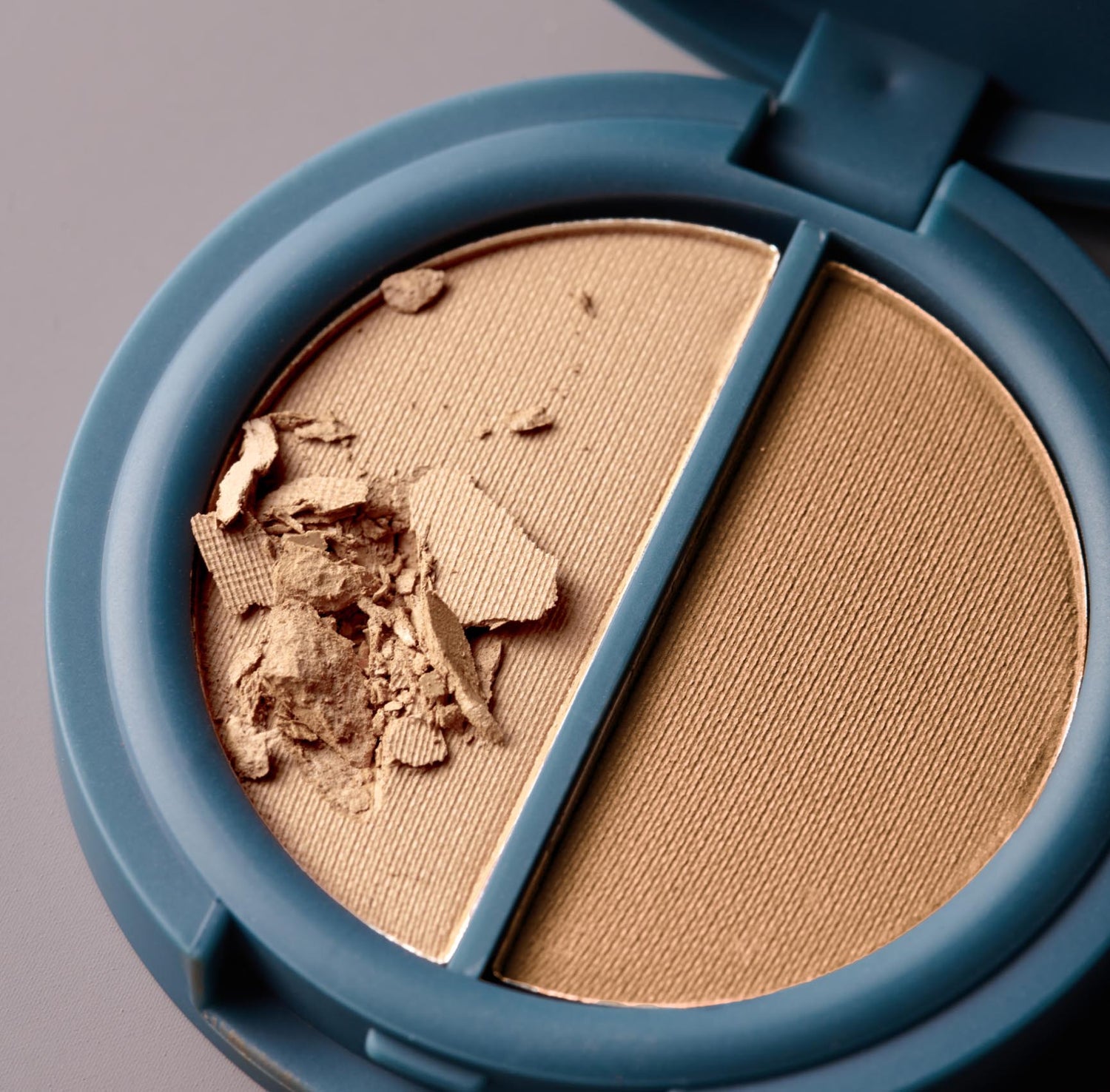 A circular, teal-colored compact of two shades of warm beige-colored pressed cosmetic powder on a gray background. 