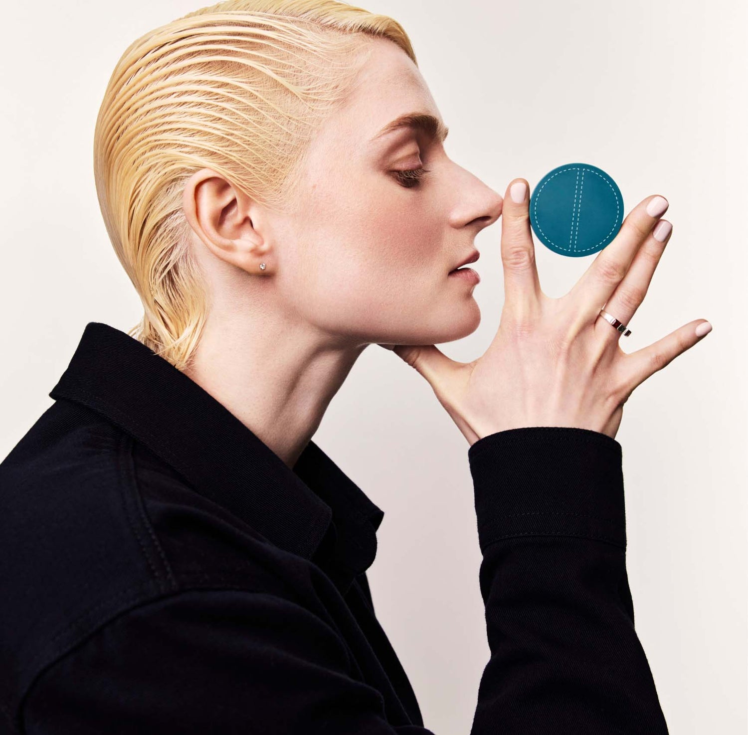 A blonde woman in a black jacket shown in profile holds a closed SPATCH invisible spot fix compact between her index and middle fingers, against a white background. The compact is teal-colored, circular, and about 2 inches in diameter. The compact has a graphic of two half-circles with dashed outlines on it.