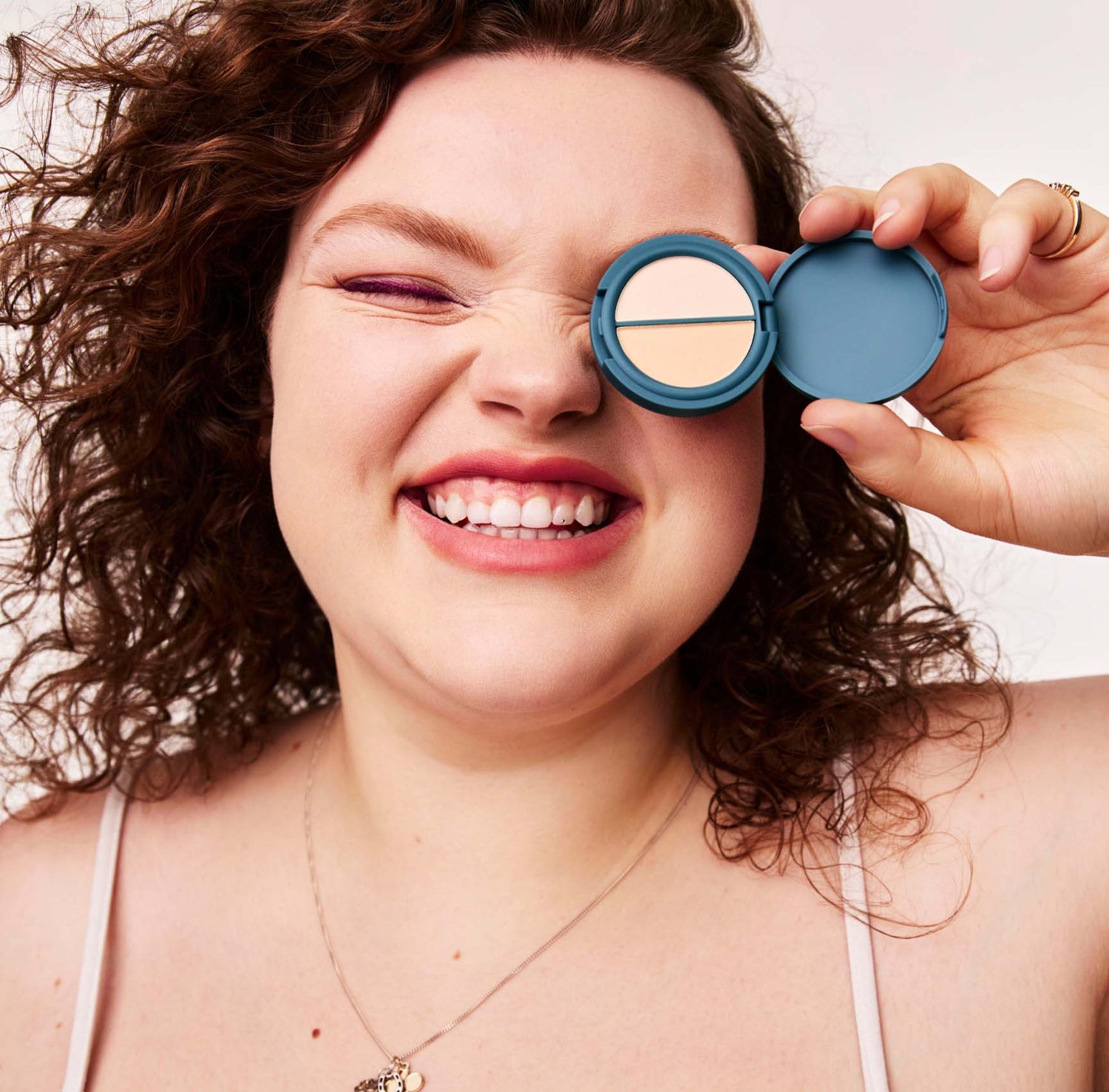 A smiling woman with curly brown hair holds an open, circular, teal-colored compact of peach-colored pressed cosmetic powder up to her eye. She is on a white background and wearing a top with white thin straps and a gold necklace.