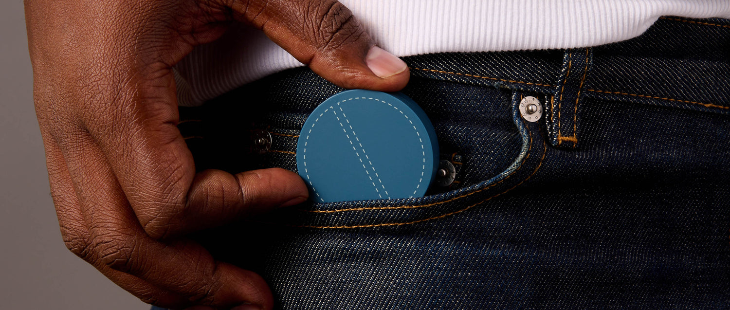 A man's hand pulls a round teal-colored cosmetic compact, about 2 inches in diameter, from a dark denim jeans pocket. A white ribbed shirt tucked into the jeans shows at the top of the photo