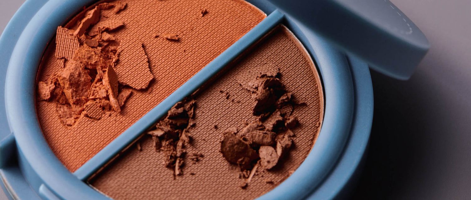 A circular, teal-colored compact of two shades of warm brown-colored pressed cosmetic powder on a gray background